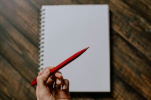 Una mano tiene una matita rossa sopra un quaderno aperto, pronta a prendere appunti.