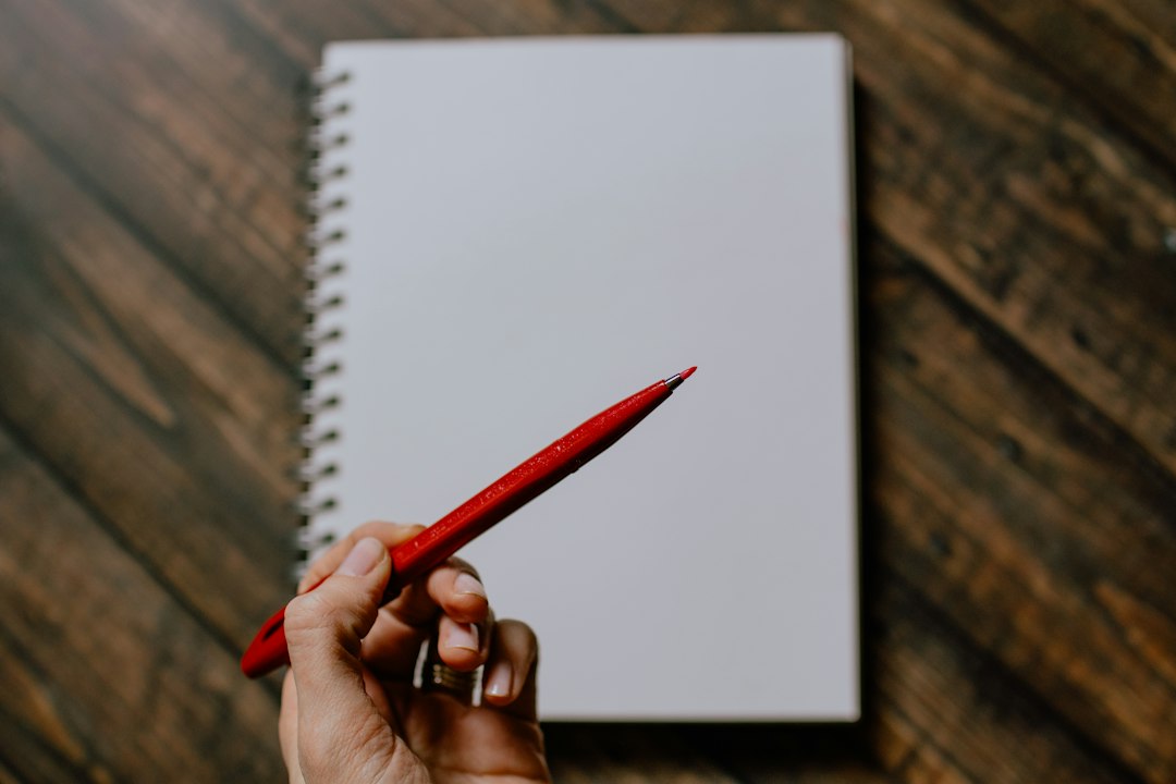 Una mano tiene una matita rossa sopra un quaderno aperto, pronta a prendere appunti.